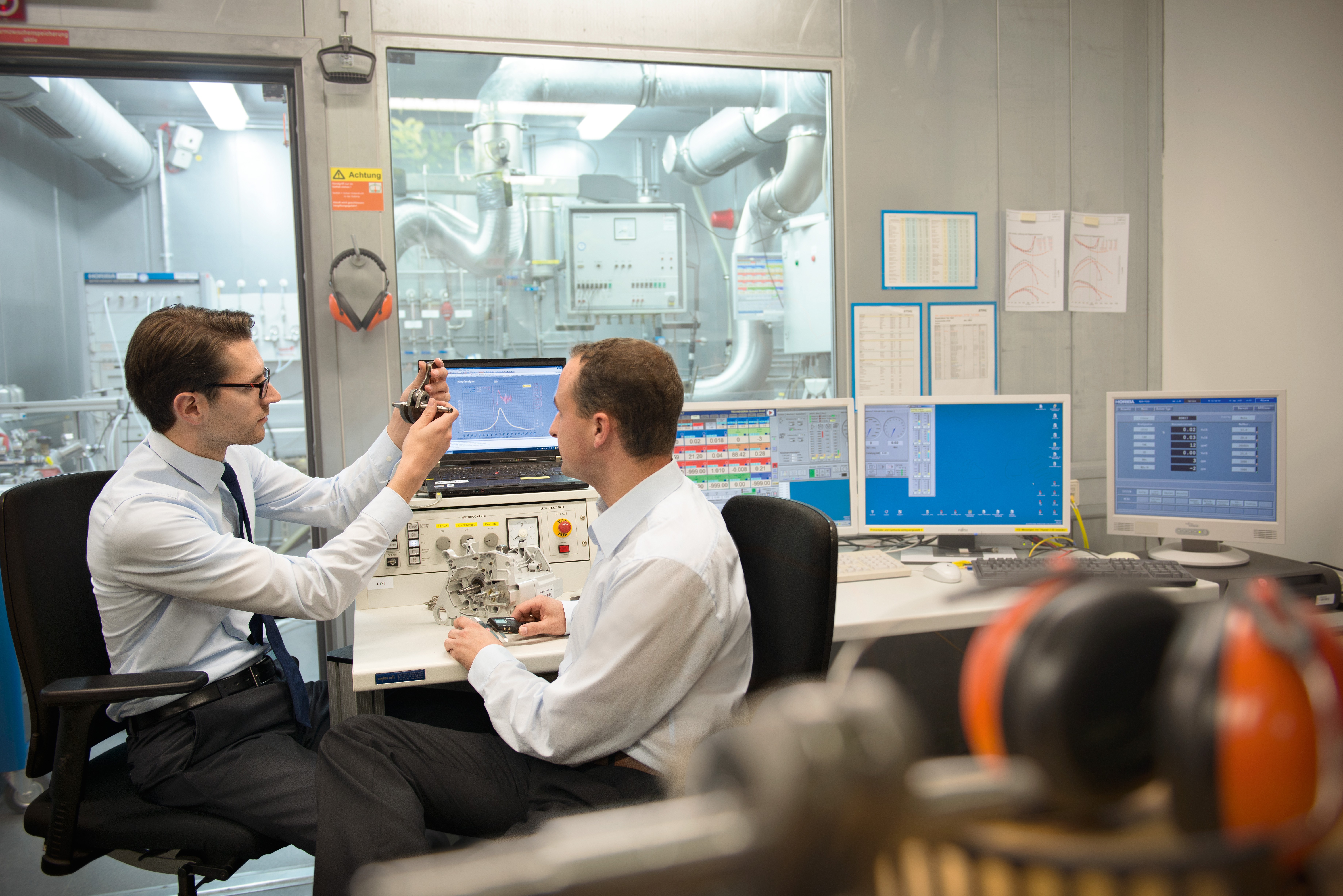 Dva muži ve výzkumu a vývoji sedí v laboratoři u stolu s několika monitory a zkoumají část výrobku.