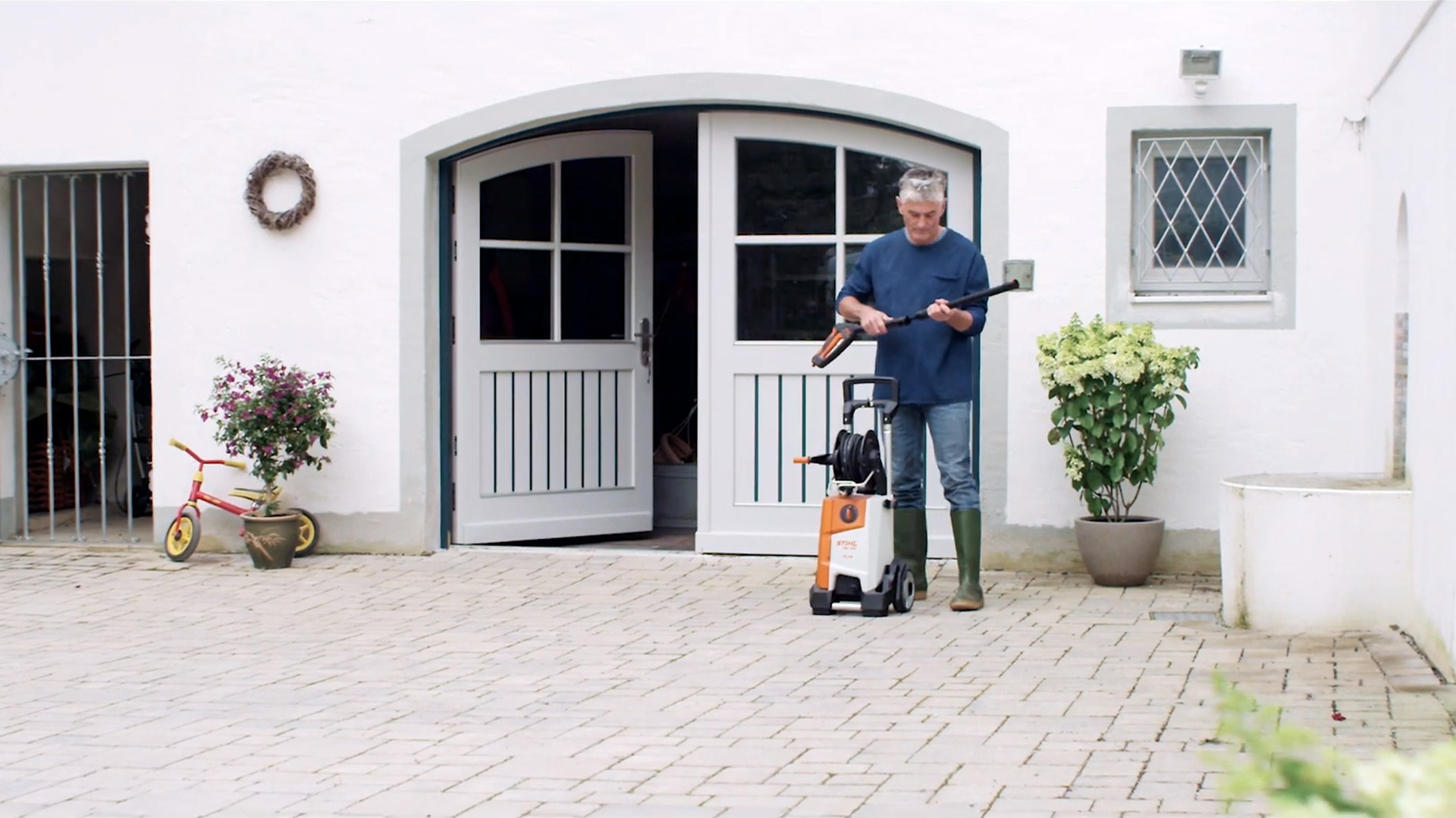 Muž stojí před bílým domem a připravuje si vysokotlaký čistič STIHL k čištění fasády