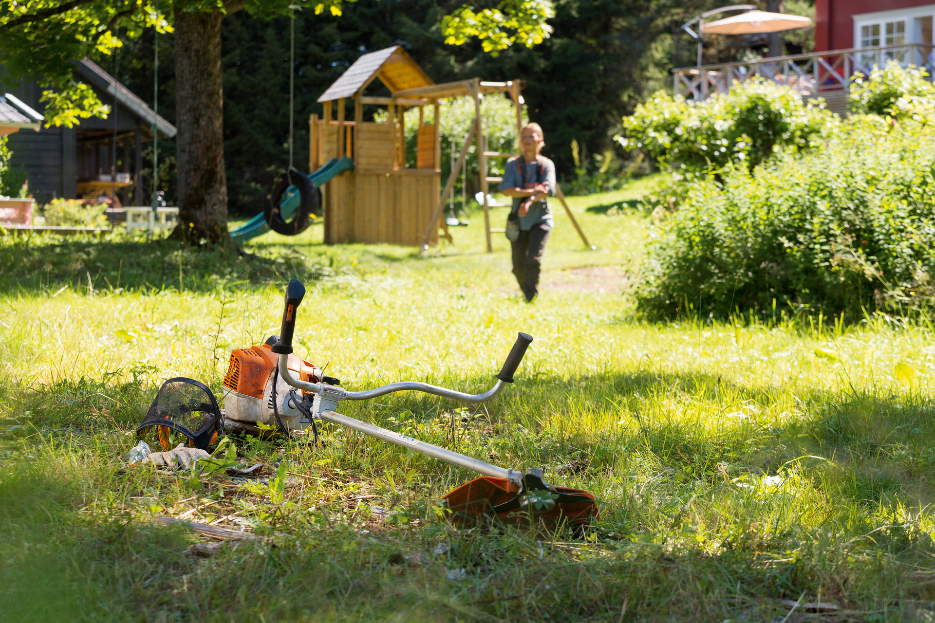 Křovinořez STIHL FS 240 leží v trávě, v pozadí se žena připravuje na jeho údržbu 