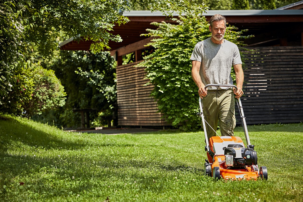 Muž při sekání trávníku s benzínovou sekačkou STIHL RM 248 před zahradním domkem.
